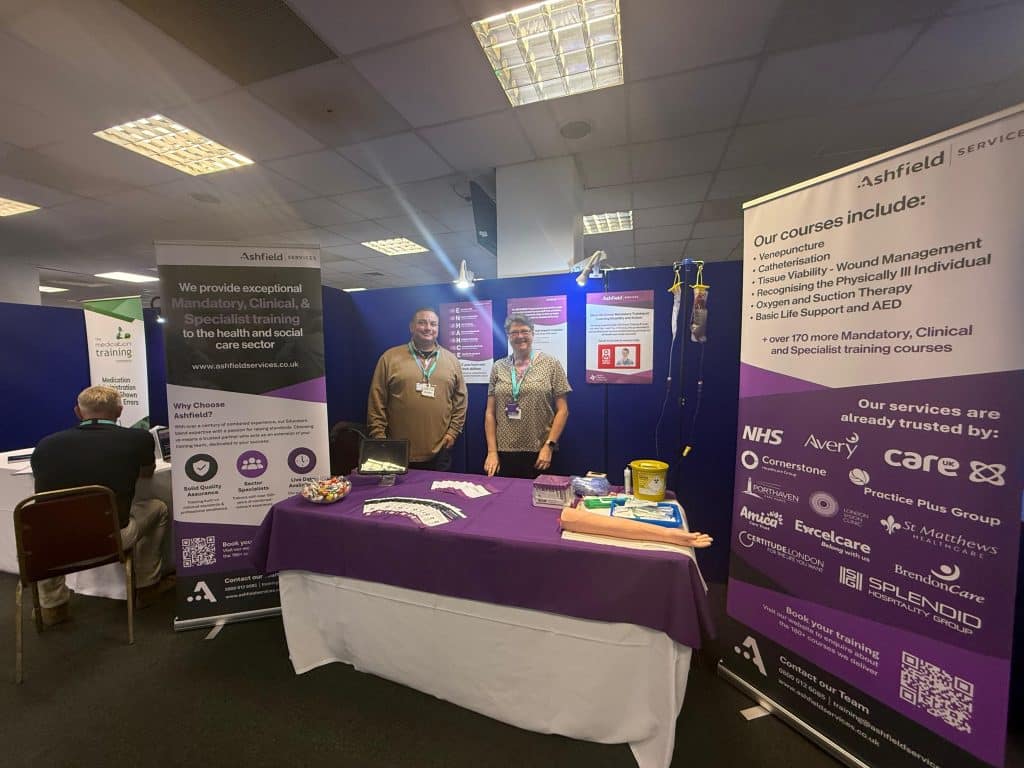 Billy and Paula from Ashfield Services standing at their exhibition stand during the Sussex Care Showcase 2025, displaying information about mandatory, clinical, and specialist training courses for the health and social care sector.