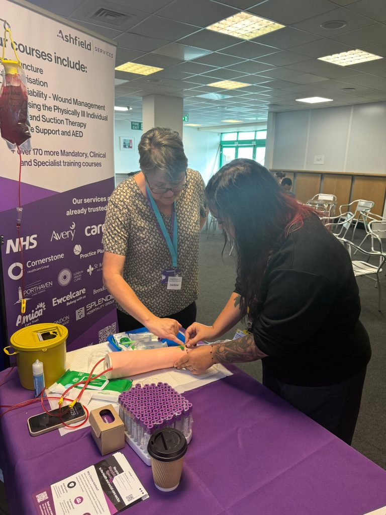 Two attendees at the Sussex Care Showcase 2025 taking part in an Ashfield Services practical training demonstration on clinical procedures, highlighting hands-on training in care.