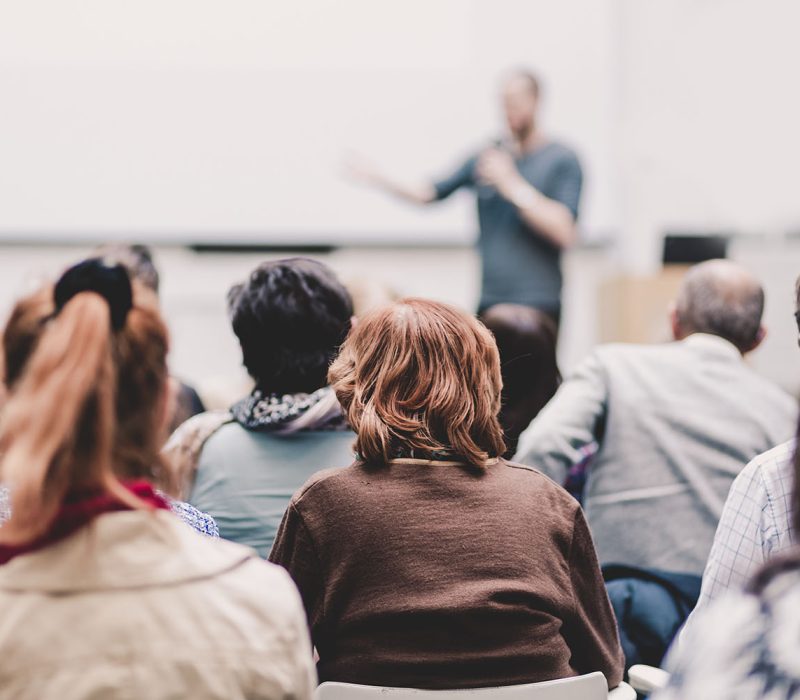 Male speaker giving presentation in lecture hall at university workshop. Audience in conference hall. Rear view of unrecognized participant in audience. Scientific conference event.