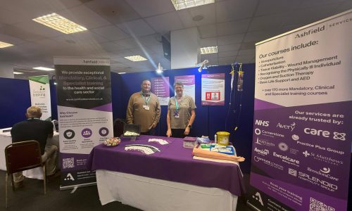 Billy and Paula from Ashfield Services standing at their exhibition stand during the Sussex Care Showcase 2025, displaying information about mandatory, clinical, and specialist training courses for the health and social care sector.